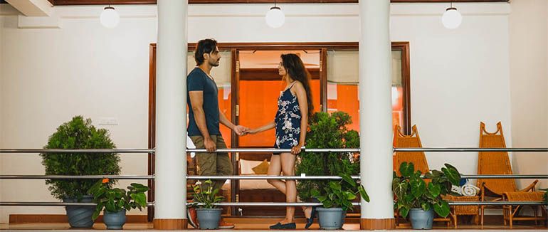 A man and woman standing on a balcony, looking at each other, surrounded by plants.