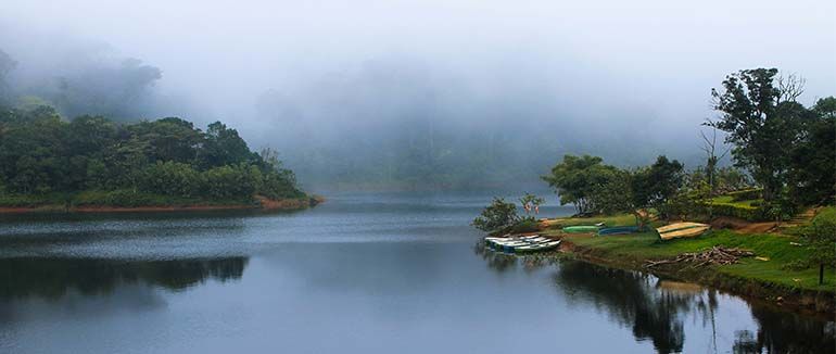 Misty lake surrounded by green hills, reflecting in calm water, with small boats on the shore.