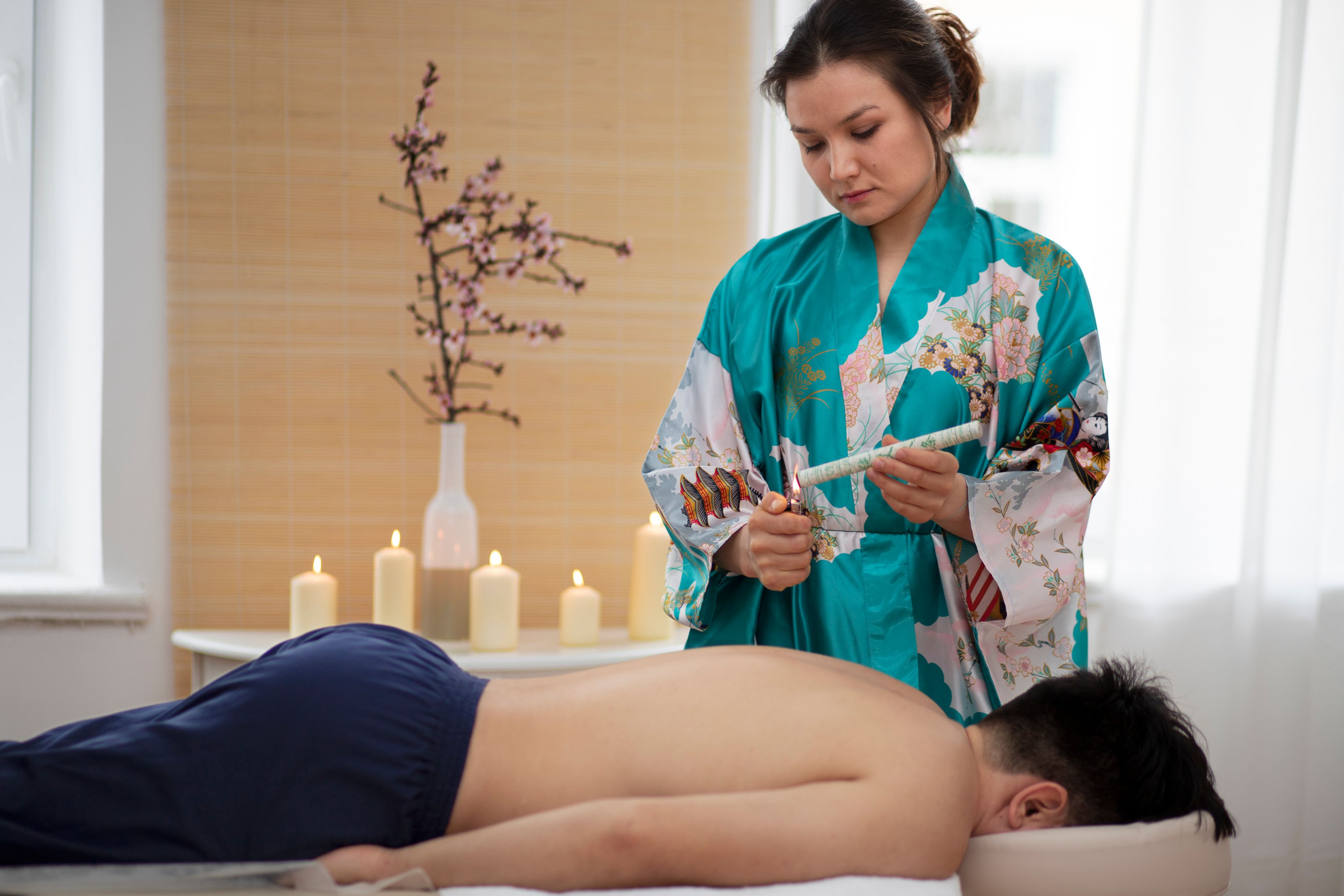 A therapist performs ear candling on a person lying face down on a massage table.