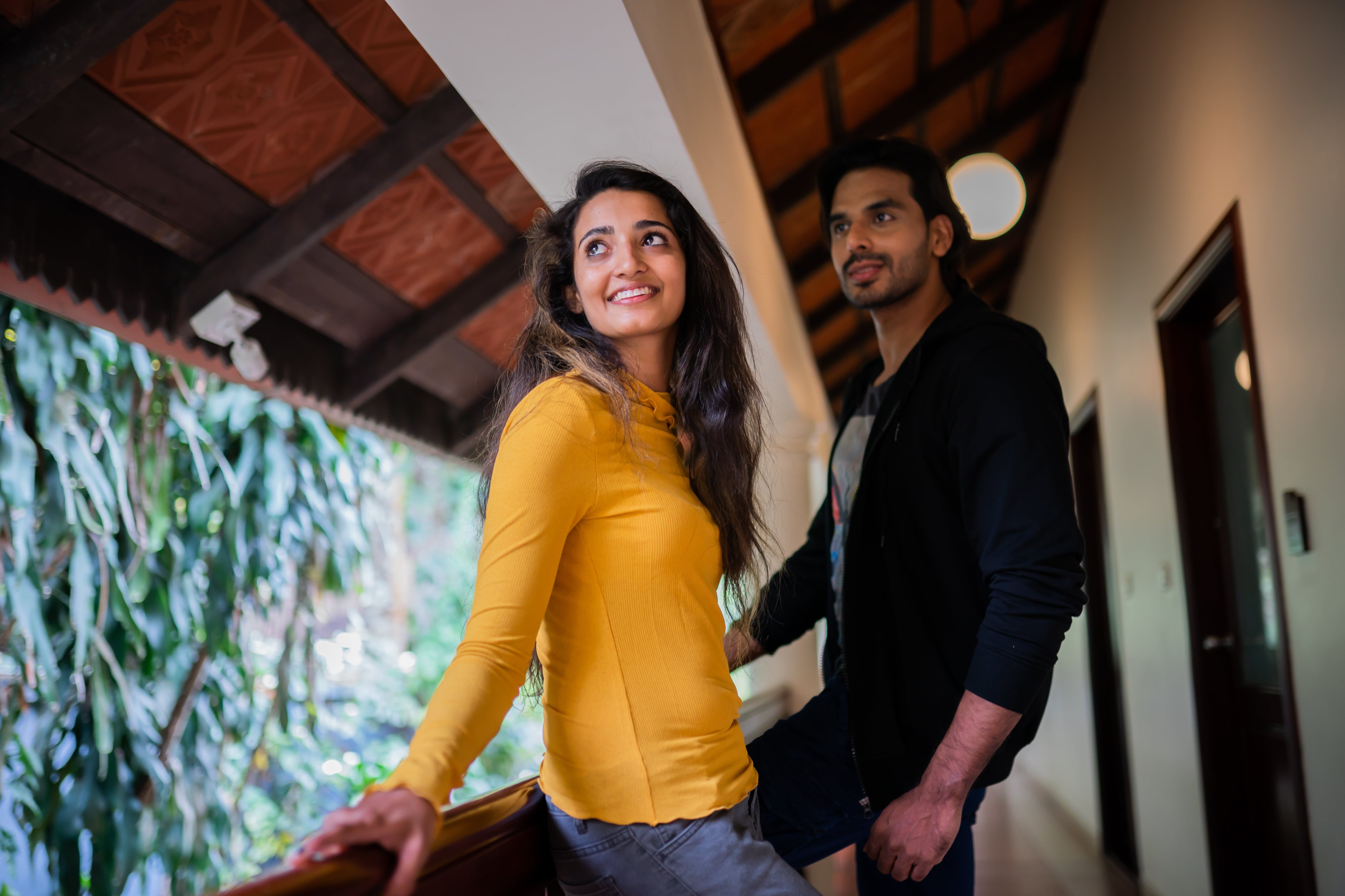 Smiling young woman in yellow with man on a rustic balcony surrounded by green plants.