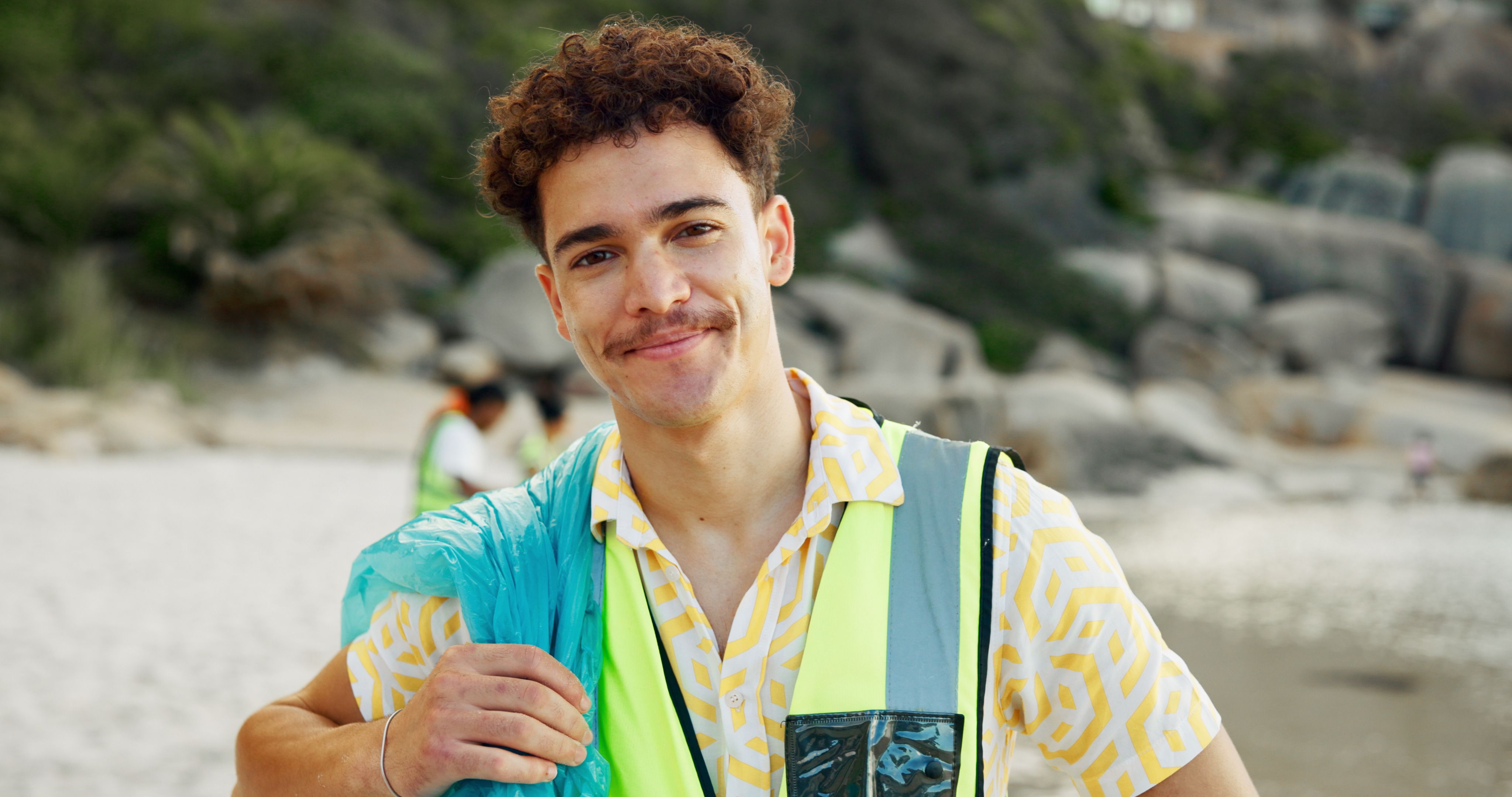 Young man in a safety vest and patterned shirt smiles on a sandy beach.