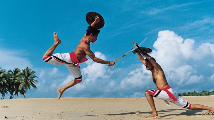 Two men perform Kalaripayattu martial arts on a sandy beach with swords and shields.