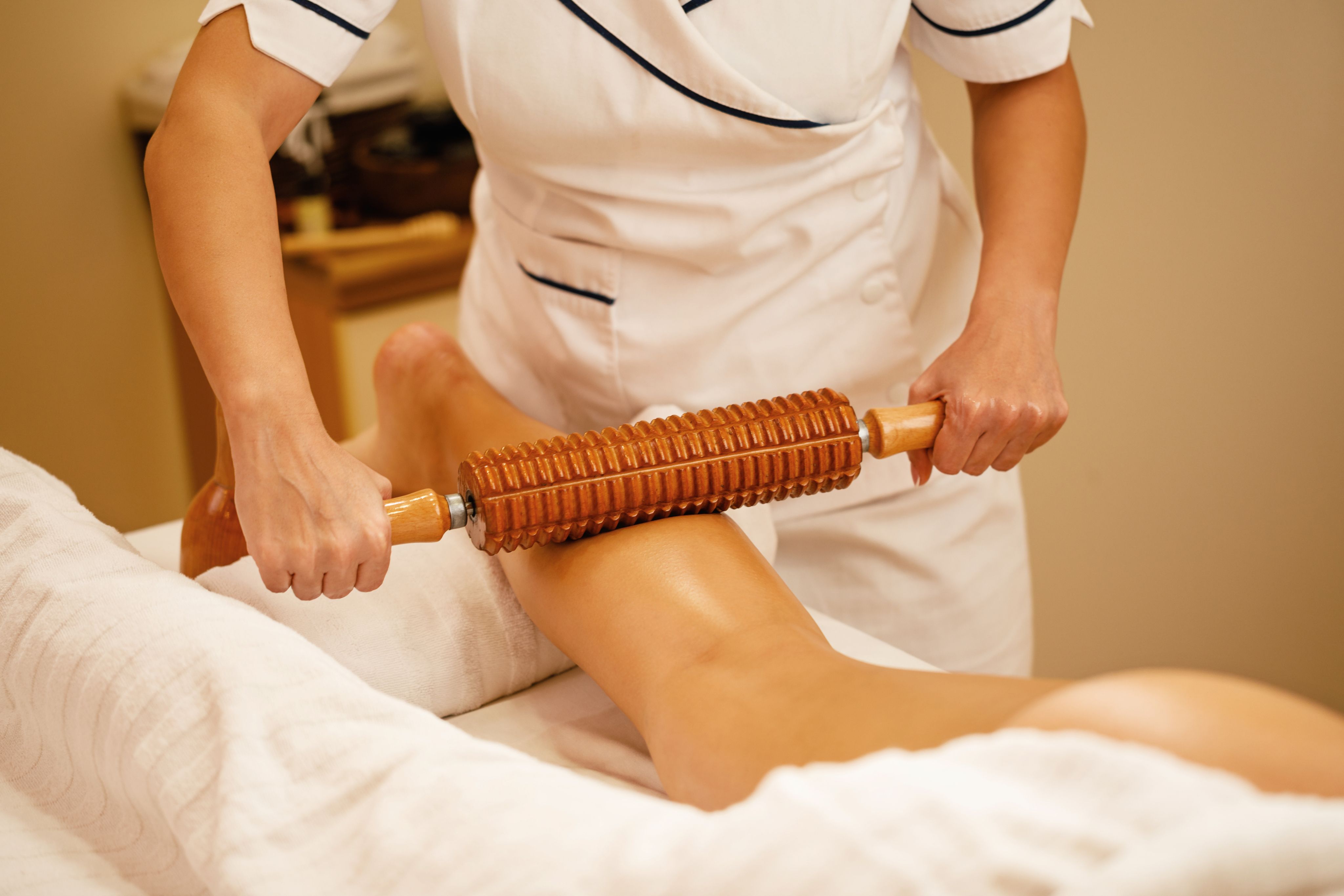 Therapist in white uniform uses a wooden roller massager on a person's bare leg.
