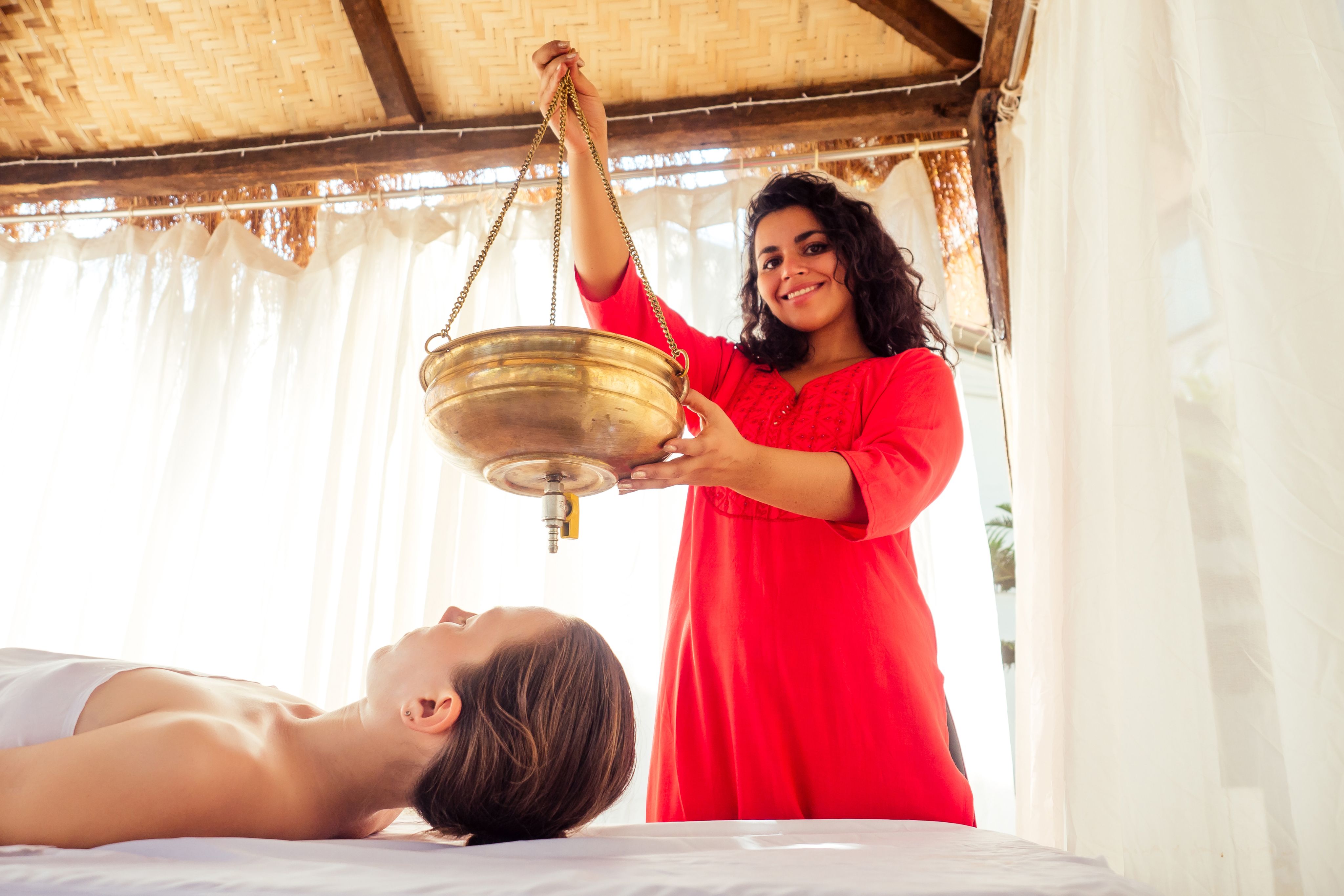 Smiling woman performs Shirodhara treatment with a brass pot on a client's forehead.