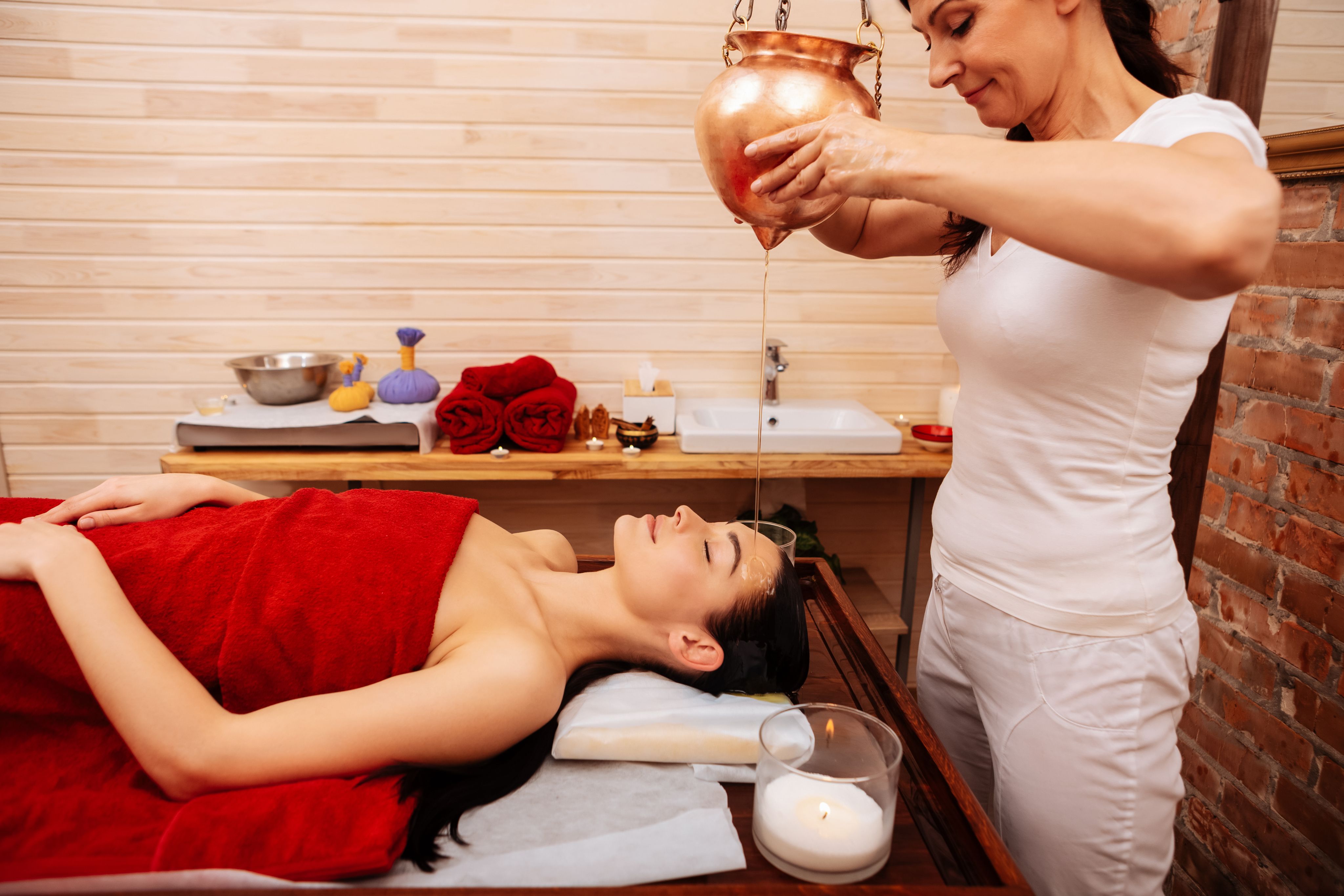 Woman receiving a shirodhara treatment, therapist pours warm oil from a copper pot.