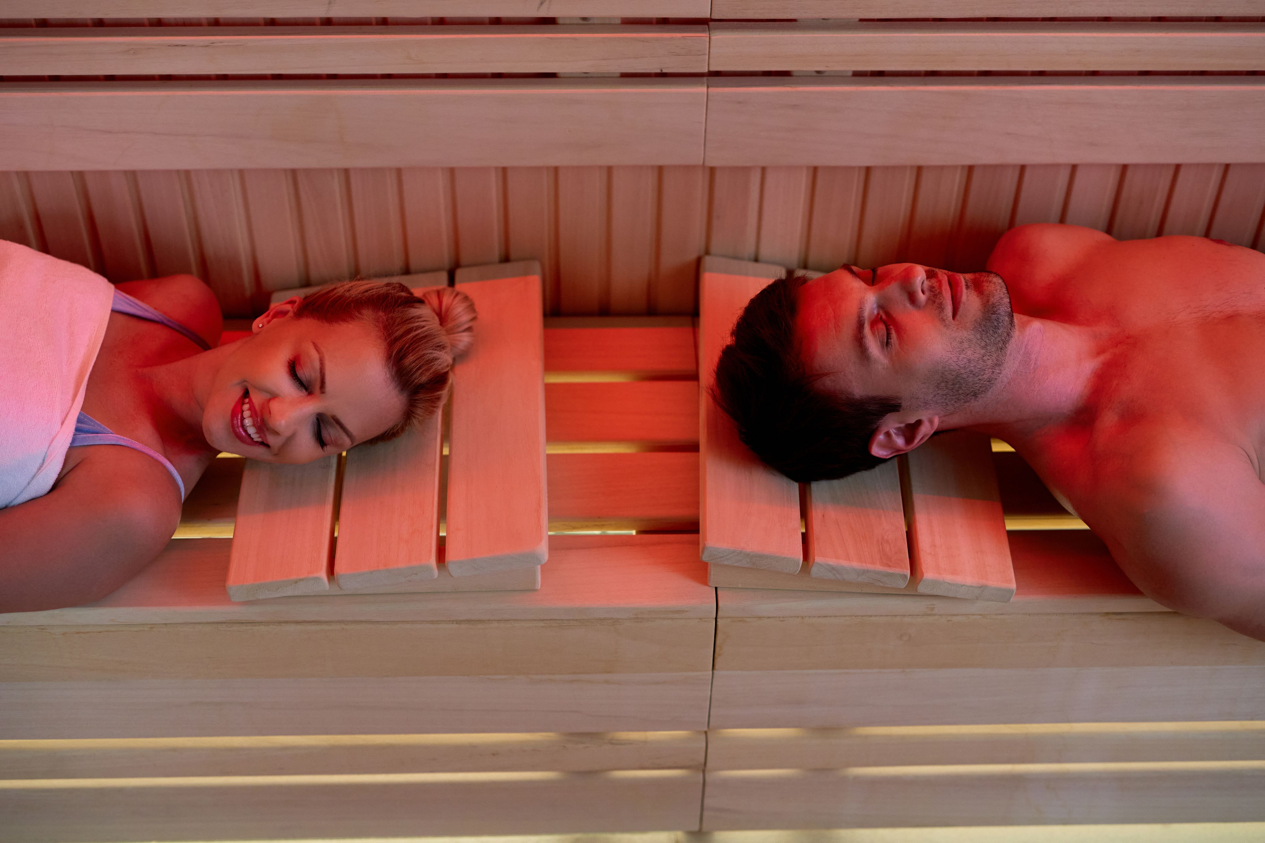 A smiling man and woman relaxing side-by-side in a red-lit infrared sauna.