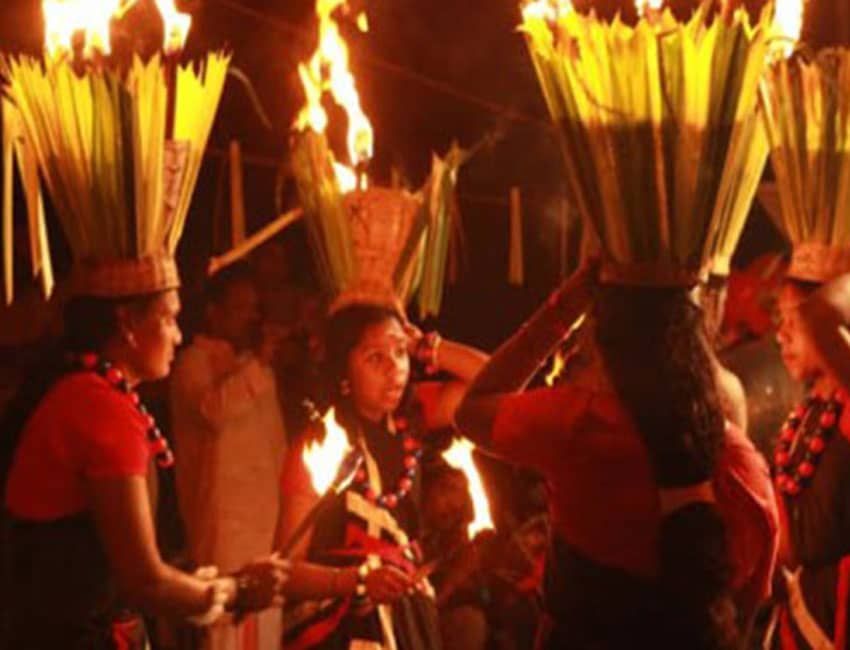 People in traditional attire with elaborate leaf headpieces celebrate amidst large flames at night.